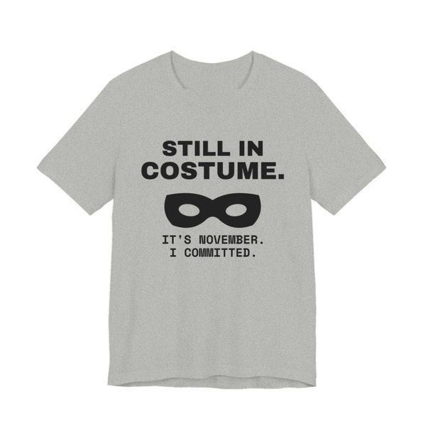 Athletic Heather t-shirt printed in dark ink with “Still in Costume.” above an eye mask, with “It's November. I committed.”