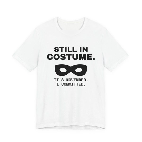White t-shirt printed in dark ink with “Still in Costume.” above an eye mask, with “It's November. I committed.”