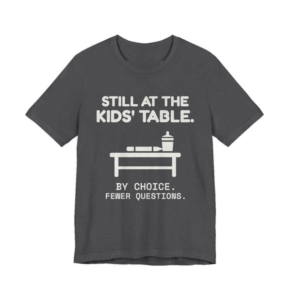 Dark Grey Heather t-shirt printed in off-white ink with the words “Still at the Kids' Table.”
